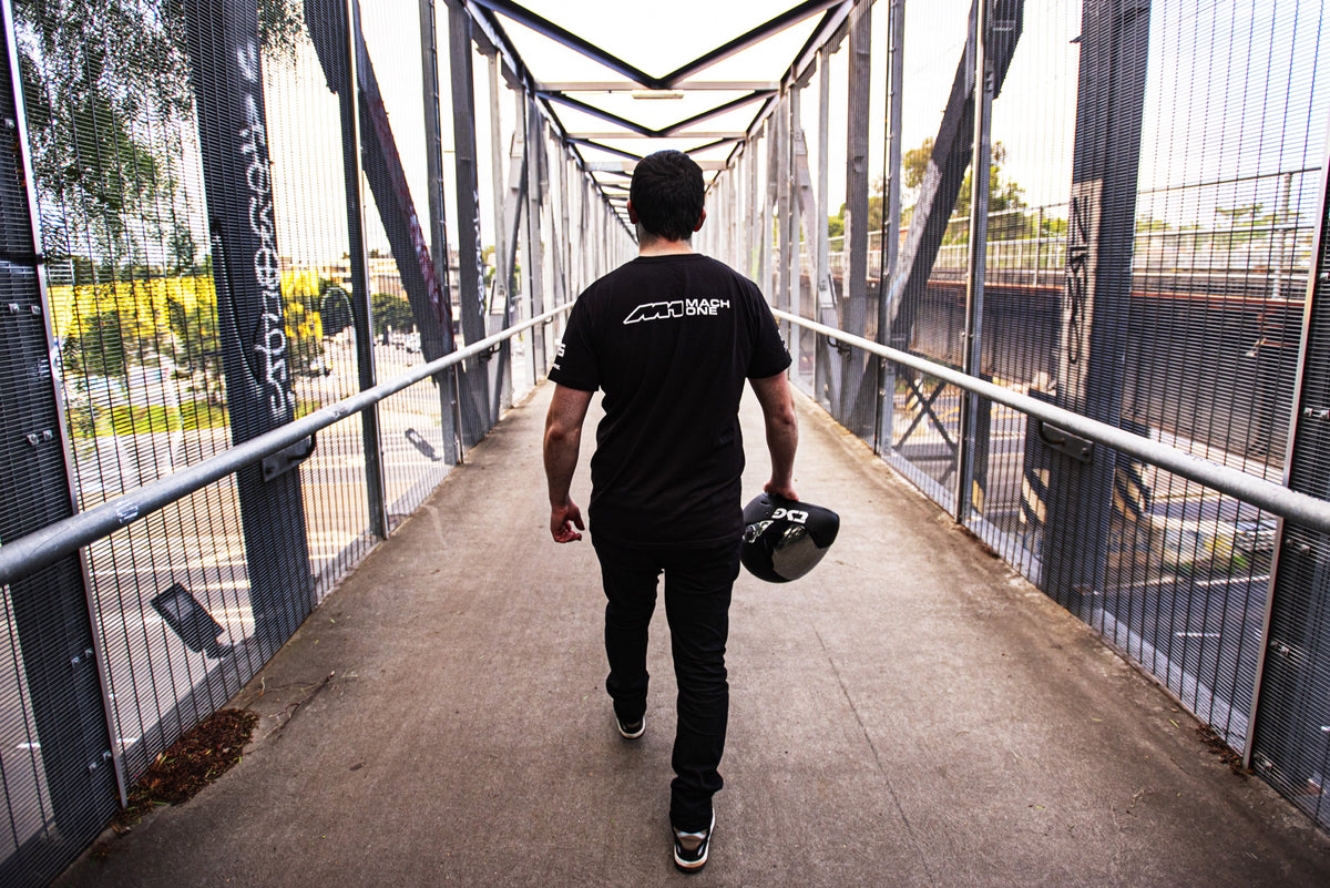 Rider walking across a bridge carrying a helmet, wearing a Mach One tee